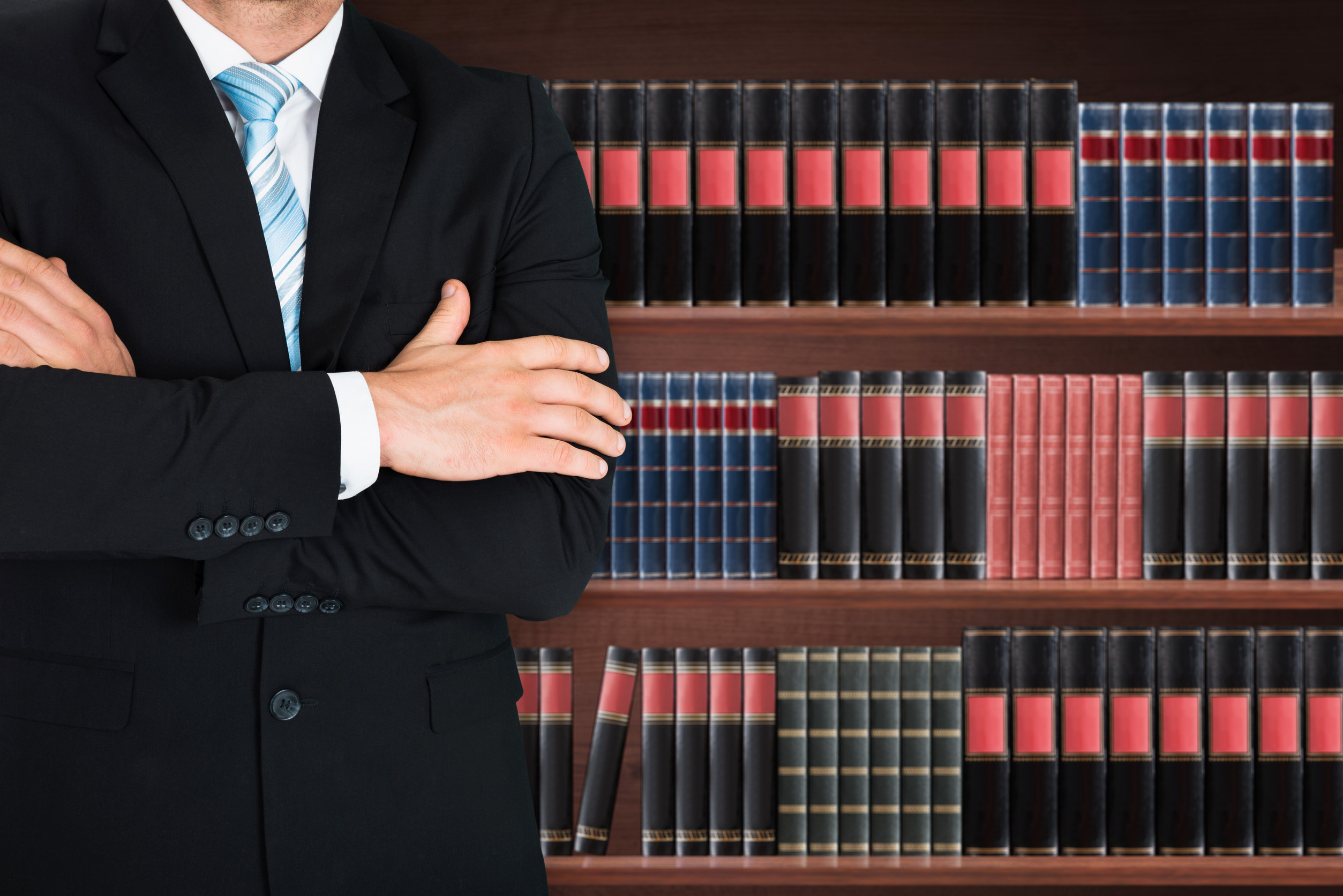 Close-up Of Male Lawyer With Arm Crossed Standing In Front Of Book Shelf